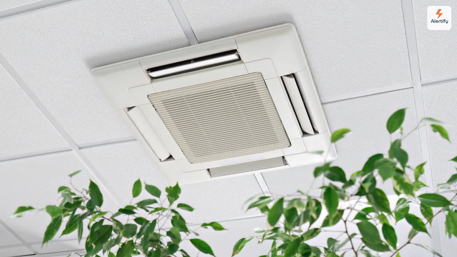 A ceiling-mounted air conditioning cassette set into a tiled ceiling, with the leaves of an indoor plant in the foreground.