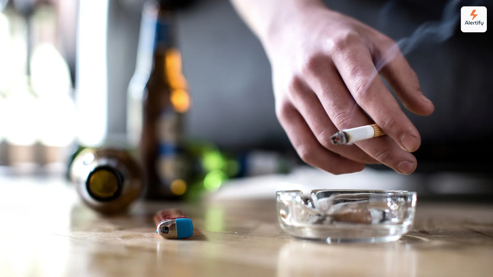 Guest holding a lit cigarette over a glass ashtray beside empty bottles on a table