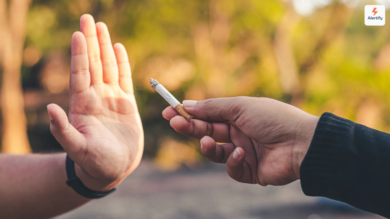 Hand raised to politely refuse a lit cigarette offered by another person outdoors