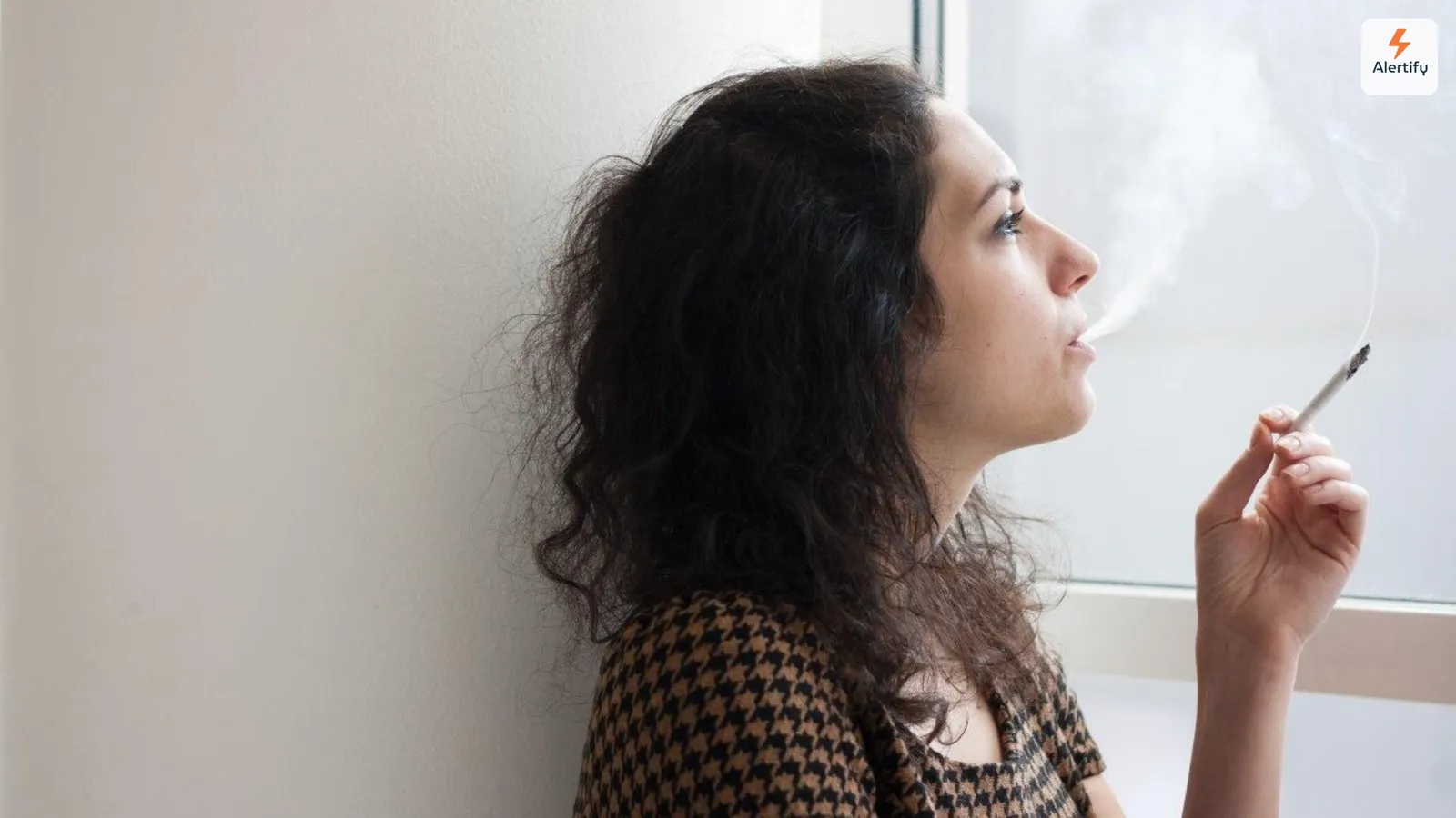 Guest leaning against a wall and smoking a cigarette indoors beside a bright window in a vacation rental